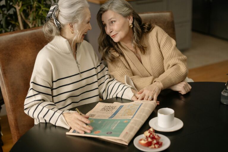 Two senior women share a moment over coffee and a crossword puzzle indoors.